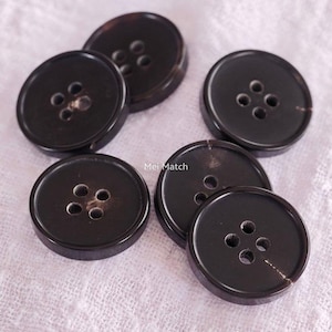 May include: Several dark brown, round buttons with four holes are displayed. The buttons have a slightly glossy finish and are scattered on a light-coloured surface. The text "Mei Match" is visible. These buttons are suitable for various sewing projects.