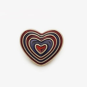 Könnte beinhalten: Eine herzförmige Brosche mit goldfarbenem Rand. Die Brosche zeigt konzentrische Herzformen in Rot, Dunkelblau und Lila. Das innerste Herz ist rot. Die Brosche ist vor einem weißen Hintergrund.