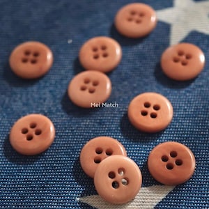 May include: A collection of ten round, peach-colored buttons, each featuring four central holes for attachment. The buttons have a smooth, glossy finish and are scattered across the frame. The text Mei Match is visible in the center.