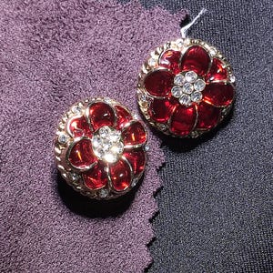 May include: Two round decorative buttons featuring a gold-tone metal frame with a central cluster of clear rhinestones surrounded by deep red enamel petals. The outer edge of each button is textured with a series of small, evenly spaced gold beads.