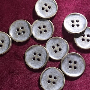 May include: Ten round metallic buttons with four holes, scattered on a deep red fabric. The buttons have a distressed, aged look with a silver-grey centre and a bronze-coloured rim. Ideal for sewing projects.