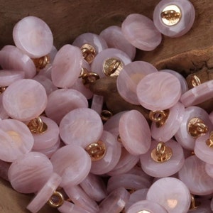 May include: A collection of pale pink buttons with a marbled effect. Each button has a gold-colored shank on the back. The buttons are round and vary slightly in size. The buttons are scattered on a wooden surface.