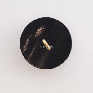 May include: A circular black button with a smooth, polished surface featuring subtle dark grey streaks. A single gold-toned metal shank loop is attached to the center of the button. The text Mei Match is printed in white letters near the center.