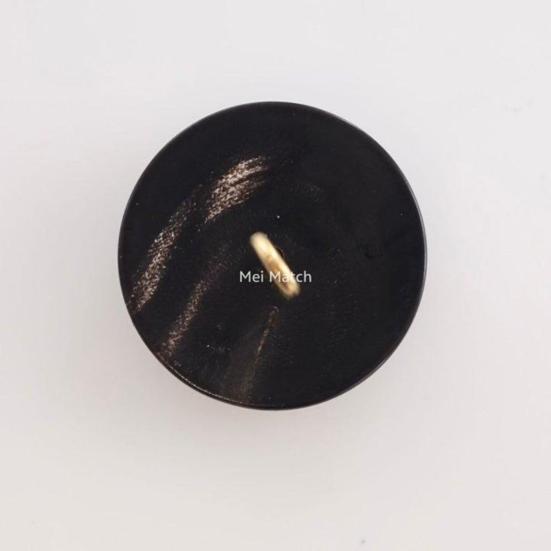 May include: A circular black button with a smooth, polished surface featuring subtle dark grey streaks. A single gold-toned metal shank loop is attached to the center of the button. The text Mei Match is printed in white letters near the center.