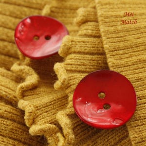 Puede incluir: Dos botones grandes, redondos y rojos con un acabado brillante están cosidos en una tela de punto de color amarillo mostaza. La tela tiene un borde rizado.