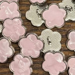 Puede incluir: Un conjunto de botones con forma de flor, con un centro rosa claro nacarado y un borde y dorso de metal plateado. Los botones están dispersos sobre una superficie de madera, mostrando su diseño decorativo.