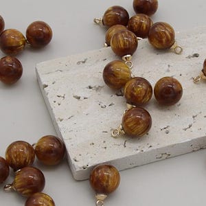 May include: A selection of spherical, brown beads with a golden-yellow pattern and gold-coloured loops. The beads are scattered on a light-coloured, textured surface. These beads are suitable for jewellery making.