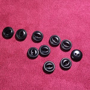 May include: A collection of black buttons with a glossy finish, scattered on a textured, deep red surface. The buttons vary in design, with some featuring an eye-shaped detail and others with two holes.