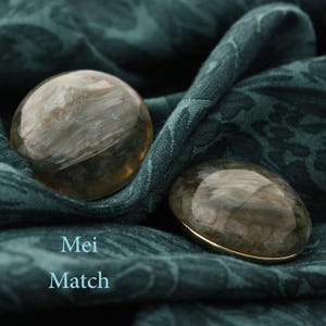 Könnte beinhalten: Zwei runde, graue und weiße Achat-Cabochons mit goldener Einfassung, auf einem dunkelgrünen Stoffhintergrund. Der Text "Mei Match" ist unter den Cabochons gedruckt.