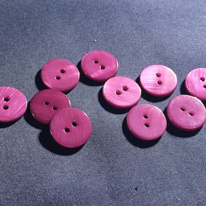 May include: Ten magenta buttons, each with two holes, scattered on a dark blue fabric. The buttons have a subtle sheen. These round buttons are suitable for sewing and craft projects. Each button is approximately 2 cm in diameter.