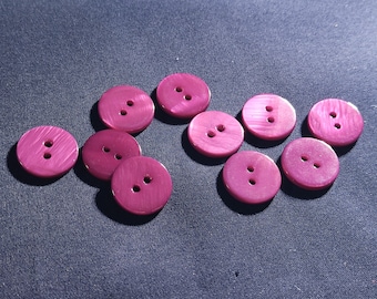 May include: A collection of ten round, magenta-colored buttons with two holes each. The buttons have a slightly iridescent sheen and are scattered on a dark blue fabric surface. These buttons are suitable for sewing and craft projects.