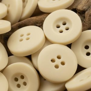 Puede incluir: Primer plano de una colección de botones de color marfil. Los botones redondos tienen dos o cuatro agujeros. La imagen muestra los botones superpuestos, con un centro ligeramente cóncavo.