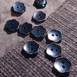 Puede incluir: Un conjunto de diez botones negros, cada uno con forma de flor de cinco pétalos y dos orificios centrales para su fijación. Los botones tienen una superficie lisa ligeramente reflectante y un borde marrón oscuro, dispuestos de forma dispersa.