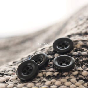May include: Three round, black, four-hole buttons resting on a textured, woven fabric with a black, white, and grey speckled pattern. The text Mei Match is visible in the center of the image, positioned between the buttons on the fabric surface.