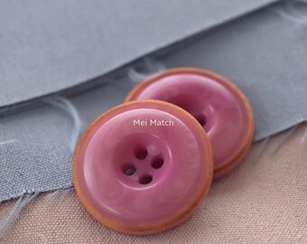 Op de afbeelding: Twee ronde knopen met een glanzend roze centrum en een beige buitenrand. Elke knoop heeft vier gaten in het midden en de tekst Mei Match in het wit op het oppervlak gedrukt. De knopen hebben een gladde, gepolijste afwerking en een licht verhoogde rand.