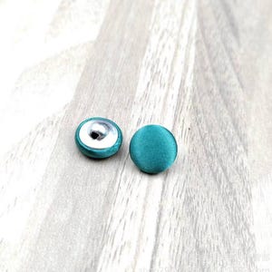 May include: Two teal fabric-covered buttons. One button has a silver metal shank on the back. The other button is solid teal. The buttons are round and have a smooth texture. They are likely for sewing or crafting projects.