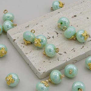 Pode incluir: Uma coleção de contas verde-claro, iridescentes, com detalhes florais dourados. Cada conta tem um pequeno laço para enfiar. As contas estão espalhadas sobre uma superfície clara e texturizada, sugerindo um tema de artesanato ou joalheria.
