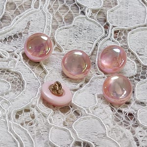 Puede incluir: Colección de botones rosas iridiscentes, incluyendo estilos redondos y planos. Los botones tienen un brillo nacarado, reflejando la luz con toques dorados y otros colores. Un botón tiene un vástago dorado.