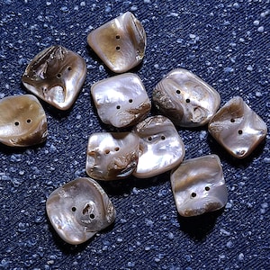 May include: A selection of iridescent shell buttons in various shapes, including squares and rounded squares. The buttons have two holes for sewing and display a mix of pearlescent white, brown, and tan hues. They are scattered on a dark blue denim surface.