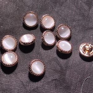 May include: A collection of nine round buttons featuring a smooth, white, cat-eye effect center inlay surrounded by a textured, metallic rose gold rim. Each button has a polished finish and a shank on the back for attachment to garments.