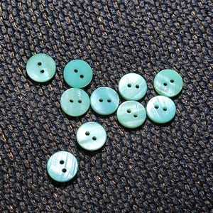 Puede incluir: Un conjunto de diez botones redondos de color verde claro, cada uno con dos agujeros, sobre una superficie oscura y texturizada. Los botones tienen un brillo nacarado. Adecuado para proyectos de costura y manualidades.