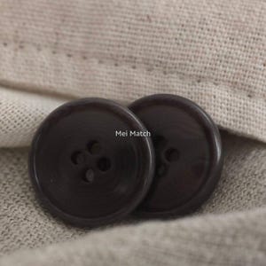 Puede incluir: Dos botones redondos de color marrón oscuro con cuatro agujeros en el centro. Los botones tienen un acabado liso y ligeramente brillante con un borde exterior elevado. El texto "Mei Match" está impreso en blanco entre ambos botones.
