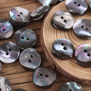 Può includere: Una collezione di bottoni iridescenti a due fori, nei toni del grigio, del nero e con sfumature di viola e verde. I bottoni sono rotondi e leggermente bombati, sparsi su una superficie di legno e una fetta di legno.