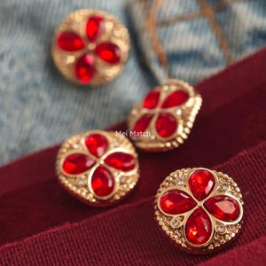 Puede incluir: Cuatro botones redondos de tono dorado con un diseño central de flor de cuatro pétalos hecho de cristales rojos en forma de lágrima, rodeados de pequeños diamantes de imitación transparentes. El texto Mei Match es visible en el centro.