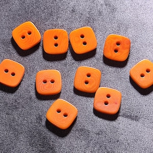 Puede incluir: Un conjunto de diez botones cuadrados de color naranja, cada uno con dos agujeros. Los botones están hechos de un material translúcido, y la superficie tiene un sutil patrón de vetas de madera. Los botones están dispersos sobre una superficie gris.