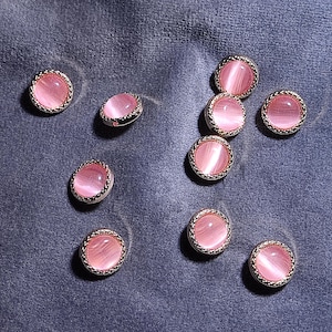 May include: A collection of nine round buttons featuring a smooth, light pink cat's eye cabochon center set within a textured, metallic silver-toned rim. Each button has a polished, reflective surface that displays a distinct horizontal light band.