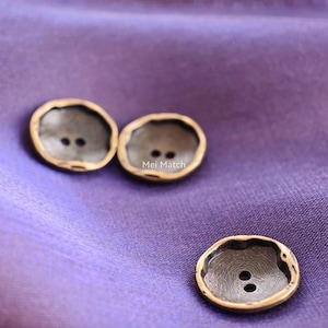 Op de afbeelding: Drie decoratieve knopen met een donker midden en een goudkleurige rand. Elke knoop heeft twee gaten om te naaien. De knopen liggen op een paarse stoffen achtergrond. De tekst "Mei Match" is zichtbaar op een van de knopen.