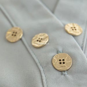 以下が含まれることがあります： 淡い青色の衣服のクローズアップ。4つの質感のある金色のボタンが付いています。各ボタンには4つの小さな穴があります。「Mei Match」の文字が1つのボタンに表示されています。ボタンは生地に縫い付けられています。