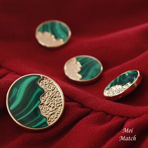 Puede incluir: Cuatro botones decorativos verdes y dorados con un diseño de mármol. Los botones están sobre un fondo de tela roja.