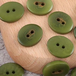 Puede incluir: Primer plano de varios botones verde oliva con dos agujeros cada uno. Los botones tienen varias formas ovaladas y redondas. Los botones están sobre una superficie de madera marrón claro.