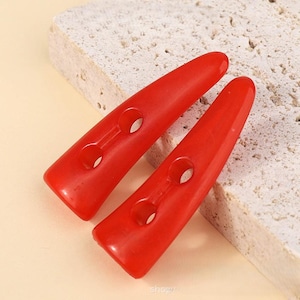Pode incluir: Dois botões de plástico vermelho em forma de chifre com dois furos cada. Os botões estão sobre uma superfície texturizada bege claro.