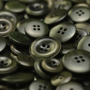 May include: A close-up of a pile of dark green buttons. The buttons are all different sizes and have four holes each.