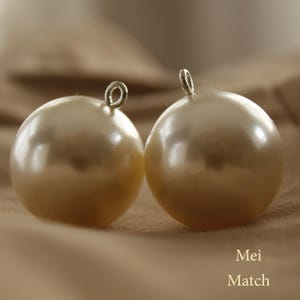 Puede incluir: Dos grandes cuentas redondas de color crema con un brillo nacarado. Cada cuenta tiene un pequeño lazo plateado en la parte superior. Las cuentas están sobre un fondo suave de tono neutro. El texto "Mei Match" es visible.