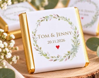 May include: A square chocolate bar wrapped in white paper with gold foil edges. The center features a watercolor botanical wreath surrounding the text TOM & JENNY, the date 20.11.2026, and a small red heart icon printed in gold lettering.