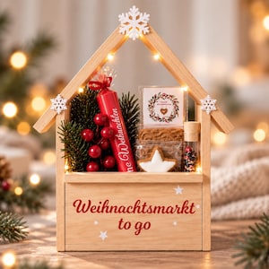 Eine helle Holzbox in Hausform mit dem roten Schriftzug "Weihnachtsmarkt to go" auf der Vorderseite. Sie enthält eine rote Kerze, grüne Tannenzweige, rote Beeren, zwei quadratische Kekse, einen sternförmigen Keks und ein kleines Glasfläschchen.