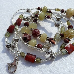 May include: A beaded lanyard featuring a variety of gemstone beads in shades of red, green, and cream. The lanyard includes faceted rectangular beads, round beads, and decorative silver-colored metal accents. A silver-colored clasp is attached.