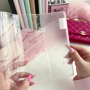 May include: A clear, pink, plastic notebook cover with a diamond grid pattern and star accents. The cover is held open, revealing the interior. The background includes a pink purse, notebooks, and a container with pens.