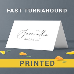 May include: A white folded card with the name "Samantha Andrews" in elegant script. The words "FAST TURNAROUND" and "PRINTED" are displayed. Scattered petals in yellow and orange are at the base of the card, on a gray background.