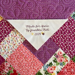 May include: A white fabric label with the text "Made for Annie By Grandma Vicki 2024" embroidered on it. The label is attached to a purple and pink patchwork quilt with floral patterns.