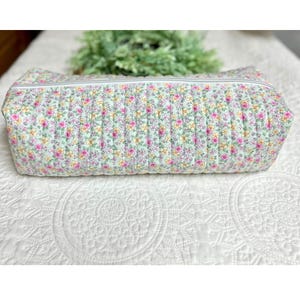 May include: A rectangular, quilted cosmetic bag with a floral pattern in pink, yellow, and white on a light green background. The bag has a white zipper and is likely made of fabric. The bag is on a white, patterned surface.