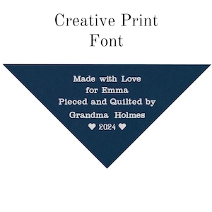 May include: A navy blue fabric triangle with white text that reads "Made with Love for Emma Pieced and Quilted by Grandma Holmes 2024" with two white hearts on either side of the year.