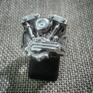 Könnte beinhalten: Ein silberner Ring mit einem detaillierten Motorrad-Motoren-Design. Der Ring befindet sich auf einem Präsentationsständer.