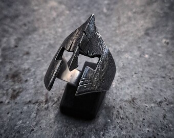 May include: A silver ring with a detailed design of a Spartan helmet. The ring is made of metal and has a textured finish.