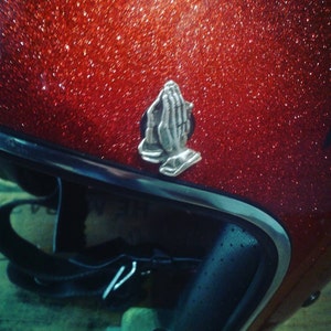 Może przedstawiać: Czerwony kask motocyklowy z srebrnym wzorem modlących się dłoni na górze. Kask ma czarną szybę i błyszczące, błyszczące wykończenie.