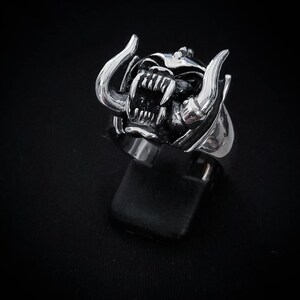 May include: A silver ring with a detailed design of a snarling animal head with horns. The ring is on a black display stand.