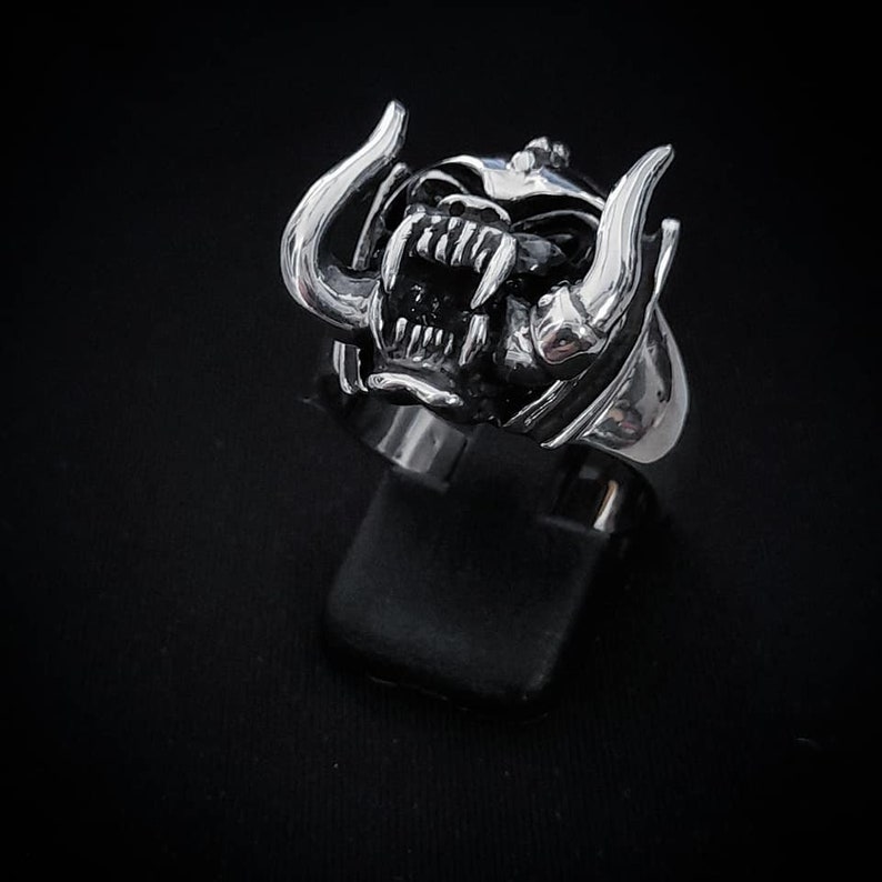 May include: A silver ring with a detailed design of a snarling animal head with horns. The ring is on a black display stand.